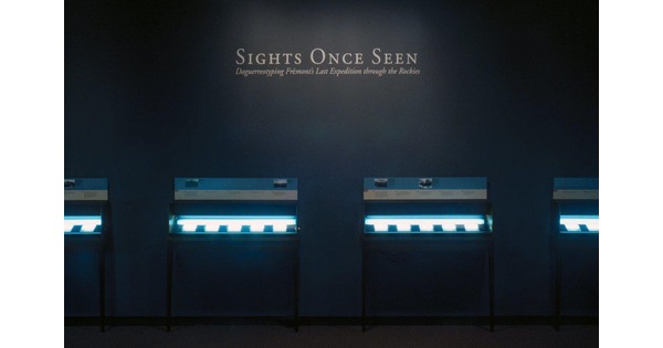 View of a dark gallery with the title spotlighted; four glass cases are lit showing objects inside.