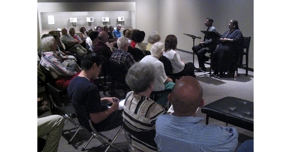 A crowd of people sit in a gallery in front of two performers who sit behind podiums.