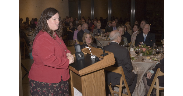 A woman stands at a podium in front of a room full of people seated at tables.