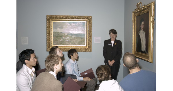 A group of adults sit in front of a painting as a docent leads a tour.