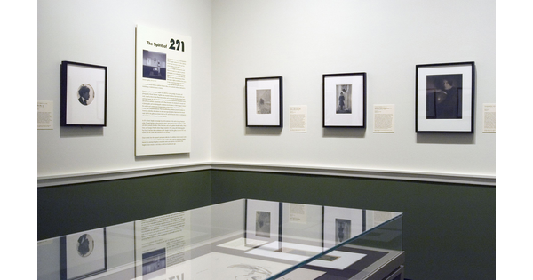 View of corner of a gallery. On the left is a framed photo and exhibition label; on the right are three framed photos; in the foreground is a glass display case containing photos.