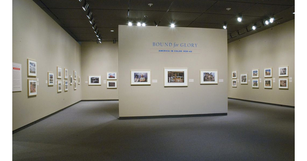 View of a gallery; the title wall features three framed photographs; more framed photos hang on the cream walls behind the title wall.