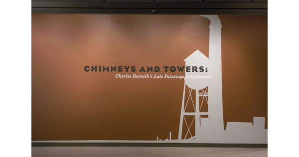 A rust-orange title wall features the title &quot;Chimneys and Towers&quot; in black over a white silhouette of an industrial chimney and tower.