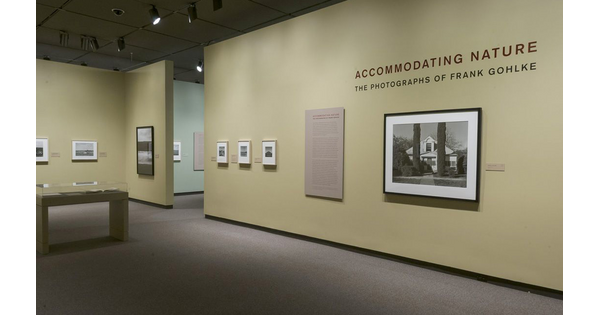 View of a gallery showing framed photographs and a text panel hanging on green walls. To the left is a display case containing several open books.