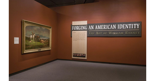 Corner of a gallery with orange walls; on the left wall is a framed painting; on the right is the exhibition title and text panel.