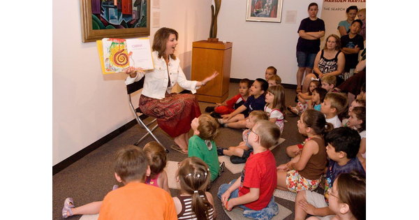 A Museum educator sits in front of an artwork and reads a story to a group of children seated on the floor. 
