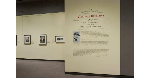 View of cream-colored gallery walls; the title wall and text is on the right; to the left one can see several framed photographs hanging on a wall.
