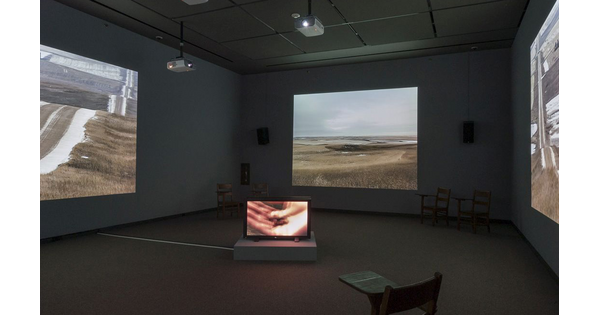 Darkened gallery with large video projections on three walls; in the center of the room is a video monitor.