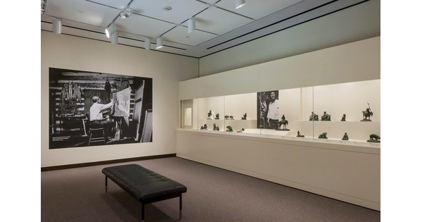 View of a gallery with a black bench in front of a display case containing many small bronze sculptures; on the wall to the left of the display case is a large black-and-white photo of a man painting in a studio.