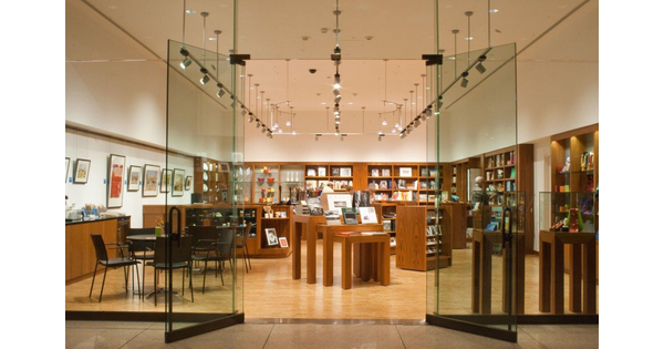 View of museum store from just outside the glass doors; wood shelves line the far and right walls and are filled with merchandise; the left wall features framed artwork; in the center are display tables and shelves featuring a variety of merchandise.