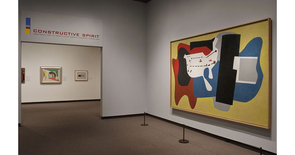 View of gallery entrance. On the right is a large abstract painting with yellow, red, blue, white, and black shapes. Through the entrance are three more paintings.
