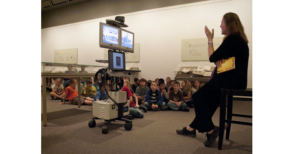 Children sit on the floor in front of a woman who is leaning on a stool and gesturing to several computer monitors in front of her.