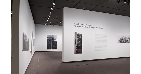 View of exhibition title wall featuring two large-scale photos mounted on a white wall; other photos can be seen on walls within the exhibition.