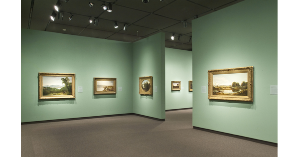 View of a gallery with light green walls. Gold-framed landscape paintings hang on each wall.
