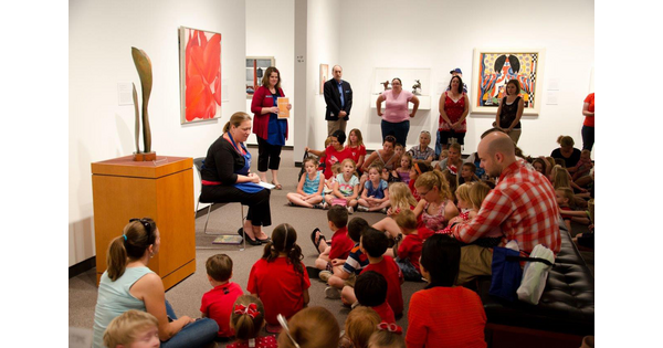A woman sits in a gallery reading a book to children and adults gathered around her.