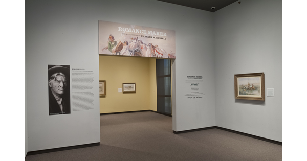 View of gallery title wall and entrance; to the left of the entrance is a large photo of the artist; over the entrance is the title graphic; on the wall to the right is a framed painting. More artwork can be seen through the entrance.