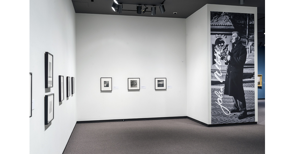 View of gallery showing five framed photographs on the left wall; three on the far wall; and a large photo of the artist to their right.
