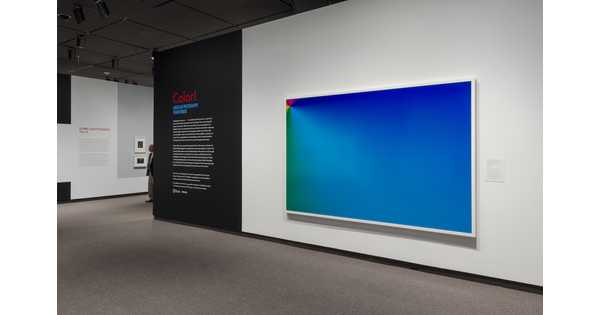 View of exhibition title wall; on the right is a large framed artwork - mostly blue with red, orange, and yellow in the upper left corner; to the left of the artwork is the exhibition title and introductory text on a black background.
