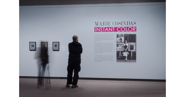 View of exhibition title wall with two people standing in front of it. On the wall are three framed photographs, the exhibition text, and an enlarged black-and-white photo of the artist.