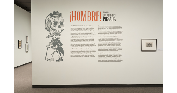 Exhibition title wall featuring a large graphic of a skeleton wearing a hat, smoking a cigar and walking with a cane; to its right is the introductory text; to the right of the text is a framed print.
