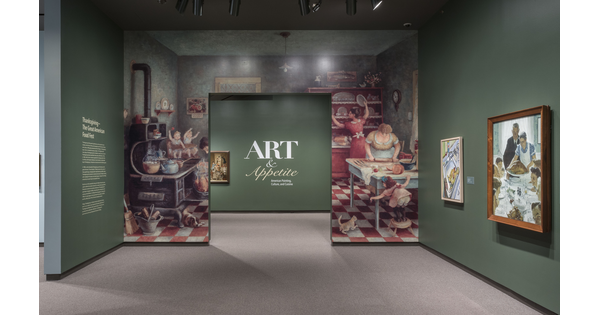 Exhibition entrance; entrance wall is covered by an enlarged illustration of people working in a kitchen; the green wall to the right features two framed artworks; through the entrance is the exhibition title.