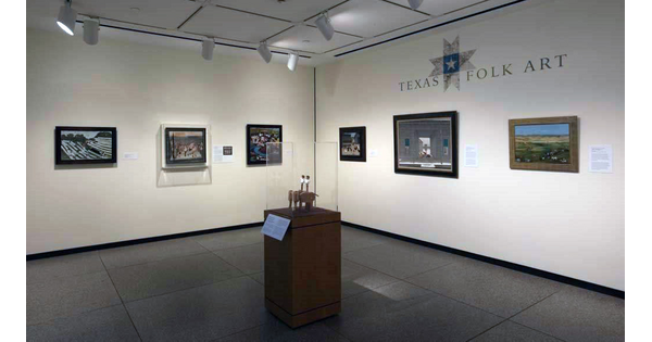 View of a gallery; both walls have three pieces of framed artwork hanging on them; above the artwork on the right wall is the exhibition title. In the foreground is a display case containing a sculpture.