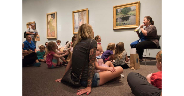 A group of children and adults sit on the floor in a gallery in front of a Carter staff member holding a book.
