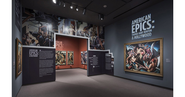 View of exhibition entrance and title wall; to the right is a large framed painting of people on a boat; the entrance wall features a reproduction of artwork showing people in an industrial setting; more framed paintings can be seen through the entrance.