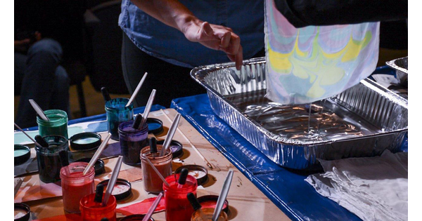 Jars of paints sit on a worktable as hands pull a piece of handmade paper out of a basin of water.