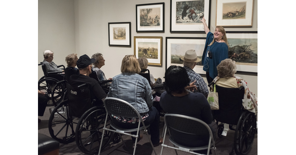 A group of senior citizens sit in chairs and wheelchairs around a woman who is gesturing toward framed artwork on a wall.
