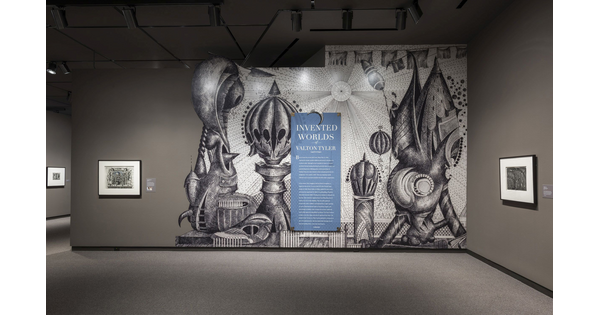 Exhibition title wall featuring an enlarged reproduction of a fantastical black-and-white drawing with introductory text on top of it. To the left and right are framed artworks hanging on taupe walls.