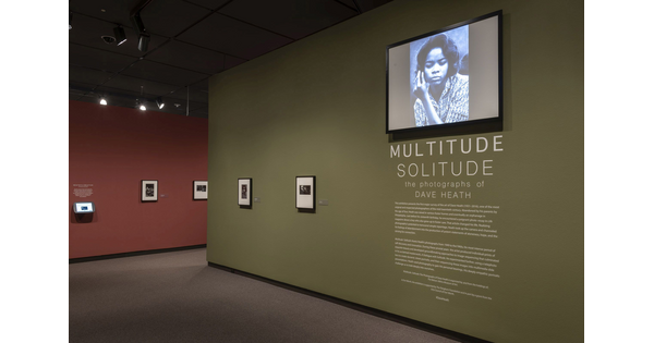 View of exhibition showing green title wall featuring two framed photographs and introductory text; above the text is a video monitor; a red wall featuring framed photos leads into the exhibition.