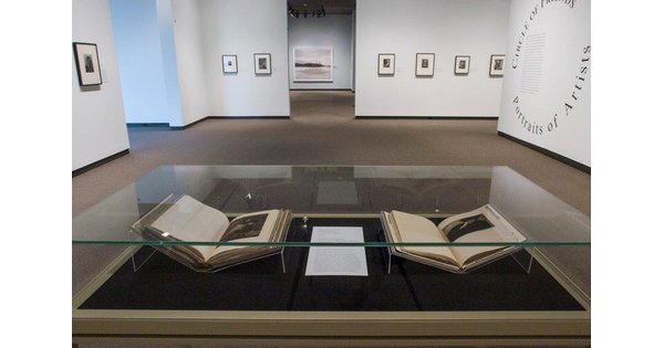 View of exhibition; in the foreground is a display case containing two open books; the title wall is to the right with the exhibition title forming a circle; on the far wall hang seven framed photographs.