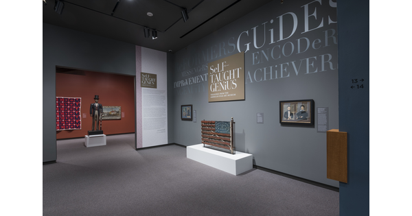 View of exhibition entrance; to the right is the title wall and graphics with two framed artworks, and an American flag made from wood; to the left is the entrance through which is a sculpture of a man in tuxedo and top hat and two works on the red wall.
