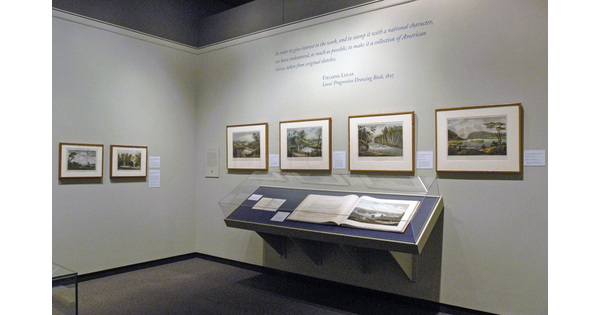 View of a corner of a gallery; two framed prints hang on a cream wall on the left, and four framed prints hand on the right. Below the four prints is a display case that contains an open book.