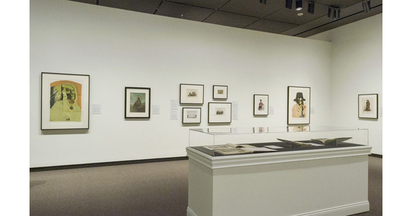 View of a gallery with white walls; framed prints of varying sizes hang on the walls. In the foreground, a glass-covered display case holds several open books.