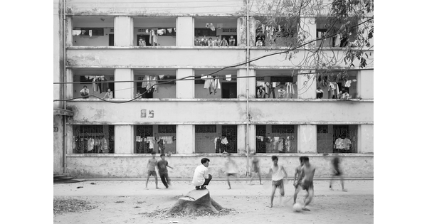 A black-and-white photograph of a group of young Asian men playing soccer in a dirt courtyard of a multi-story apartment building.