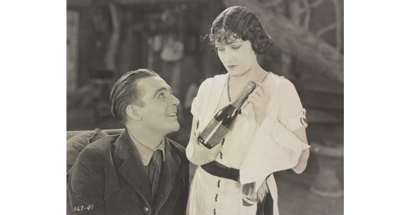 Black and white photo of a seated man looking up at a woman standing next to him holding a bottle of wine