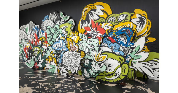 An installation photo of large wood cut-outs of flowers and leaves, some brightly-colored, against a black wall with shadows projected on the floor in a Carter gallery. 