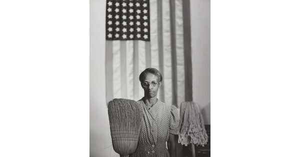 A black-and-white photograph of a middle-aged Black woman wearing a housedress, an American flag hanging behind her, and an upturned broom and mop in front of her.