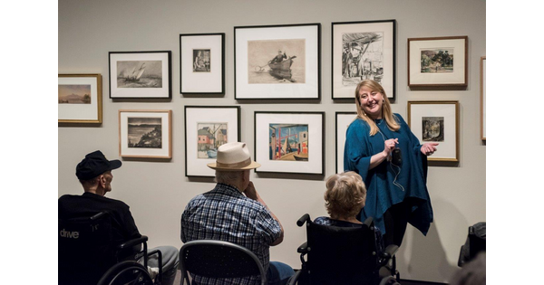 A Carter educator stands in front of a wall of framed art talking to elderly people about the artwork.