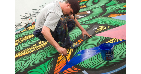 A White man squatting down to apply blue paint to an abstract mural.