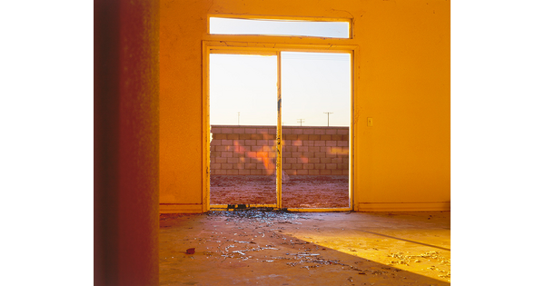 A color photograph looking through the sliding doors, one with a broken window, of a yellow room out onto a brick wall.