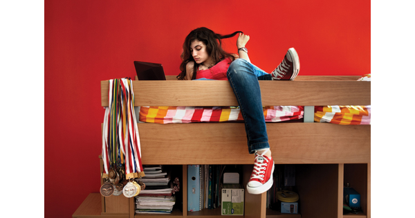 A color photograph of a light-skinned teenage girl lounging on a top bunk, twirling her long hair around a finger, sports medals slung around a bedpost.