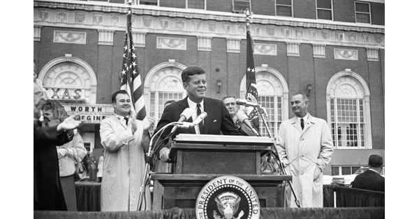 A black-and-white photograph of President John F. Kennedy speaking at a podium in front of a brick building.