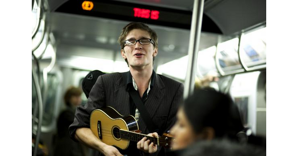 A color photograph of a White man with glasses playing a ukulele and singing inside of a subway train.