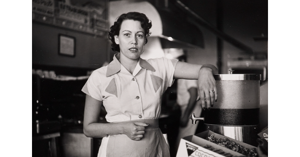 A black-and-white photograph of a White woman in a waitress uniform standing behind a diner counter with an arm propped on drink dispenser.