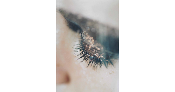 A close-up color photograph of a White woman&#039;s eyelid covered with sparkly eyeshadow and her lashes thick with mascara.