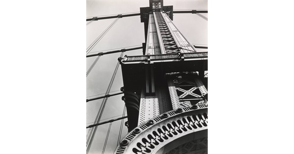 A black-and-white abstract photograph of a partial view of the tower of a suspension bridge from the ground.