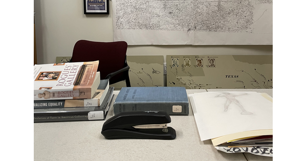 An office desk with stacked library books, manilla file folders, and a black Swingline stapler.